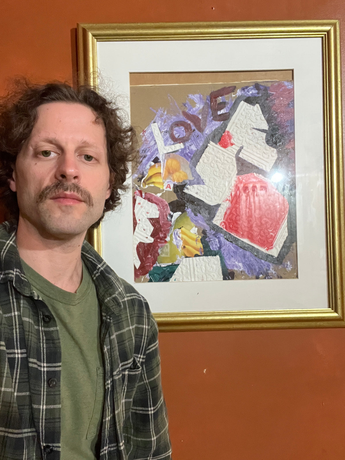 A man wearing a green shirt and a dark plaid flannel shirt leans in close to a framed abstract painting hanging on a terracotta-colored wall.